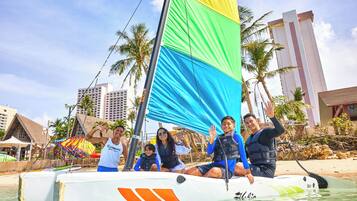 A sailboat with a colorful sail, people on board, and a beach with palm trees and buildings in the background.