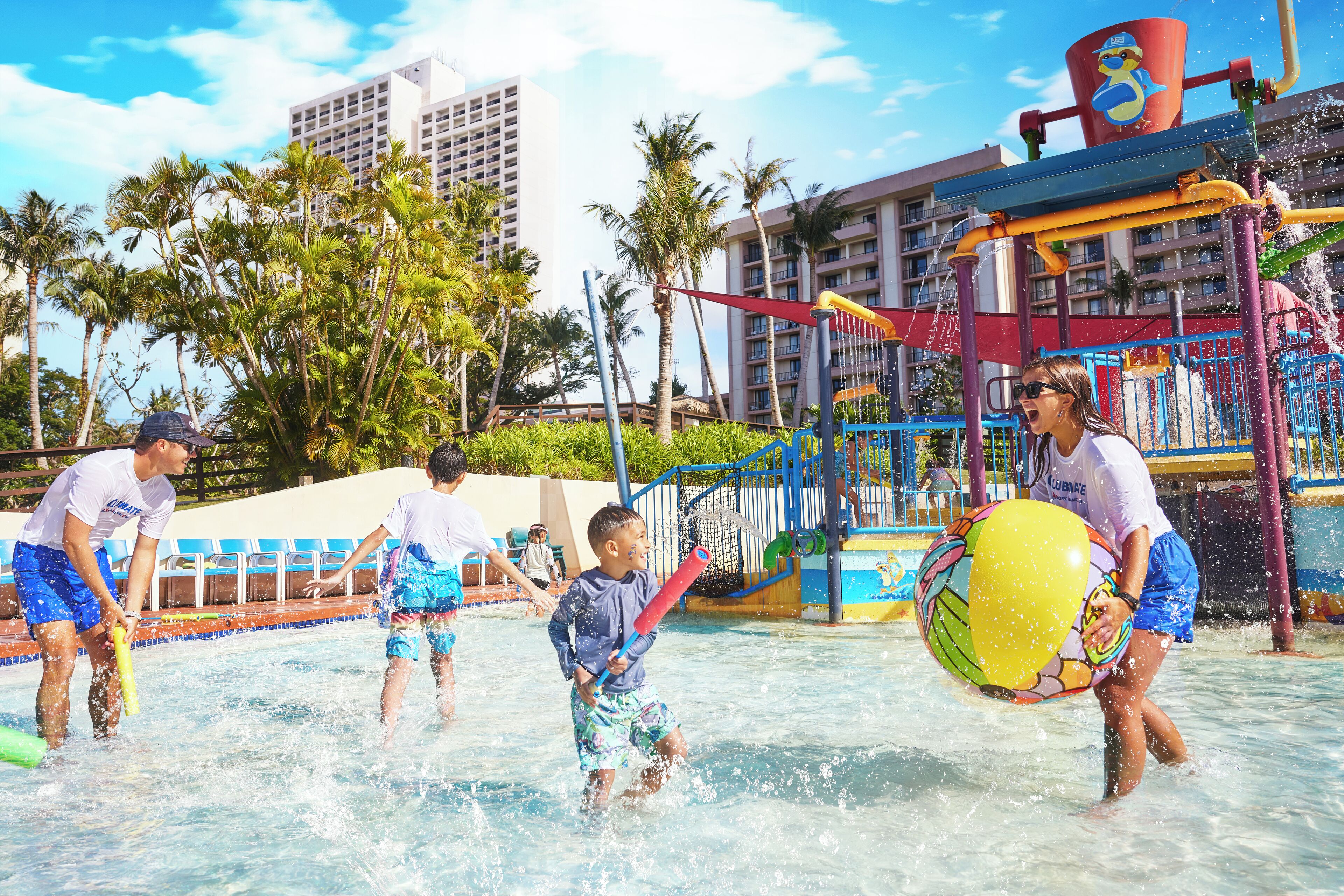 A water park with a play structure, palm trees, and a building in the background.