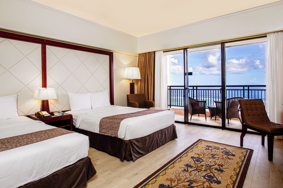 A hotel room with two beds, a sitting area with a chair, a table, and a view of the ocean through large windows.