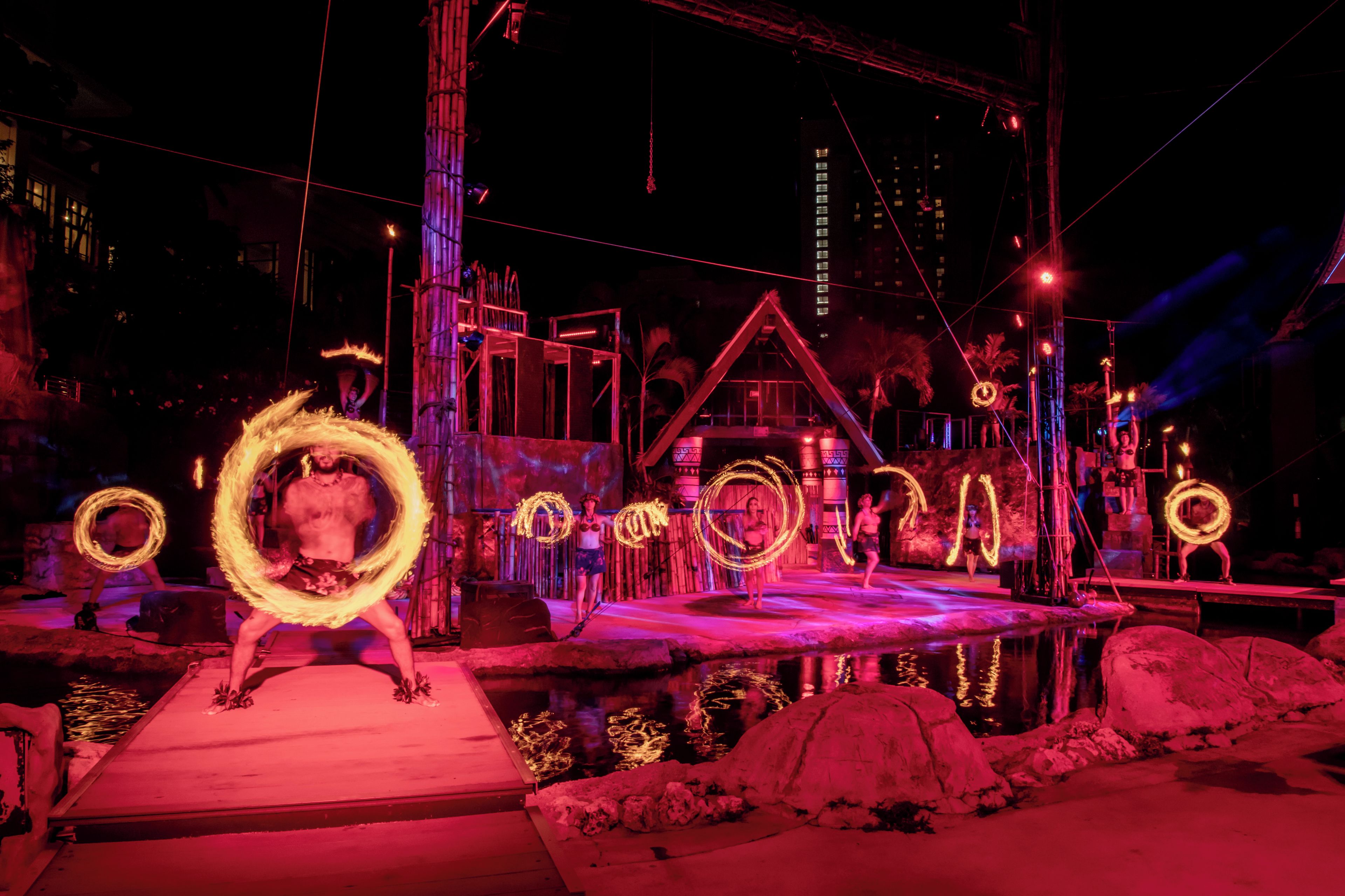A nighttime outdoor performance with performers using fire hoops, a stage with a backdrop of buildings, and a water feature in the foreground.