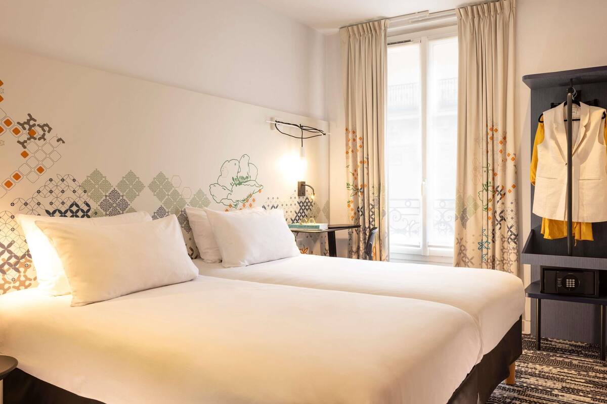 Une chambre d’hôtel avec deux lits, une petite table avec une lampe, une fenêtre avec des rideaux et un porte-manteau avec une veste.
