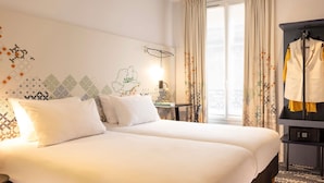Ein Hotelzimmer mit zwei Betten, einem kleinen Tisch mit Lampe, einem Fenster mit Vorhängen und einem Kleiderhaken mit Jacke.