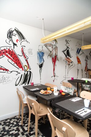 Un comedor con piso a cuadros blanco y negro, sillas de madera y mesas con superficie negra. Se observan obras de arte en las paredes que representan figuras abstractas y un lámpara colgante.