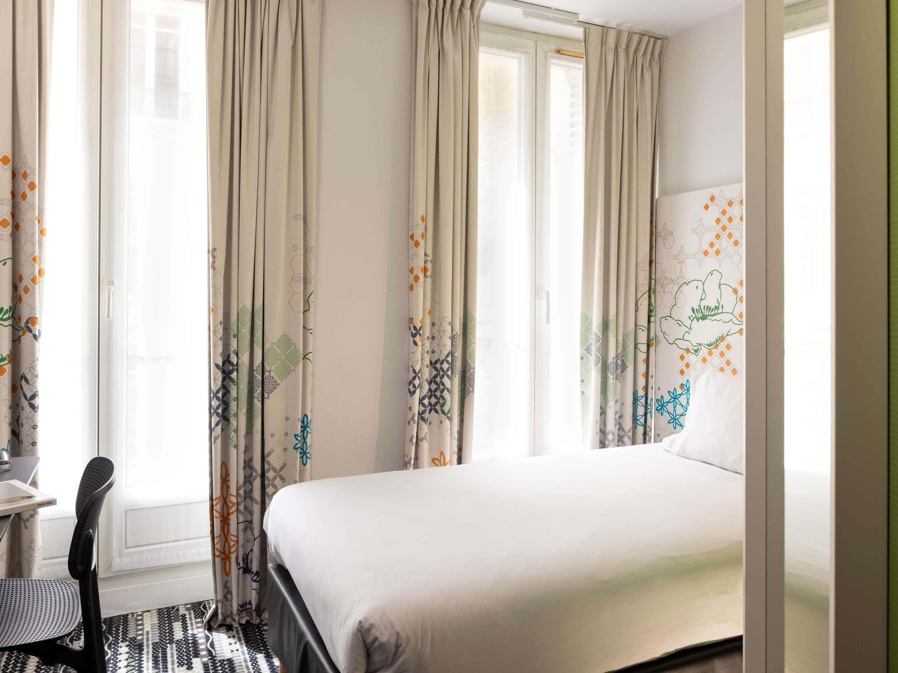 Une chambre d’hôtel comprenant un lit, un bureau, une chaise et des rideaux à motifs géométriques.