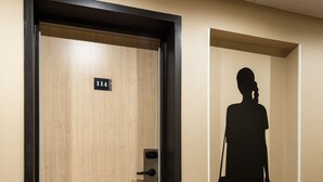 Ein Hotelzimmer mit einer Holztür, einem rot-weiß gestreiften Teppich und einer schwarzen Silhouette einer Frau an der Wand.