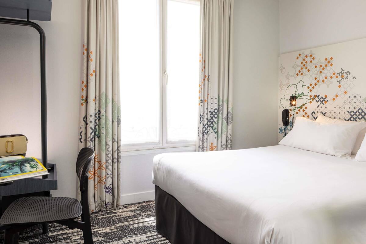 Une chambre d’hôtel avec un lit, un bureau, une chaise et une fenêtre avec des rideaux.
