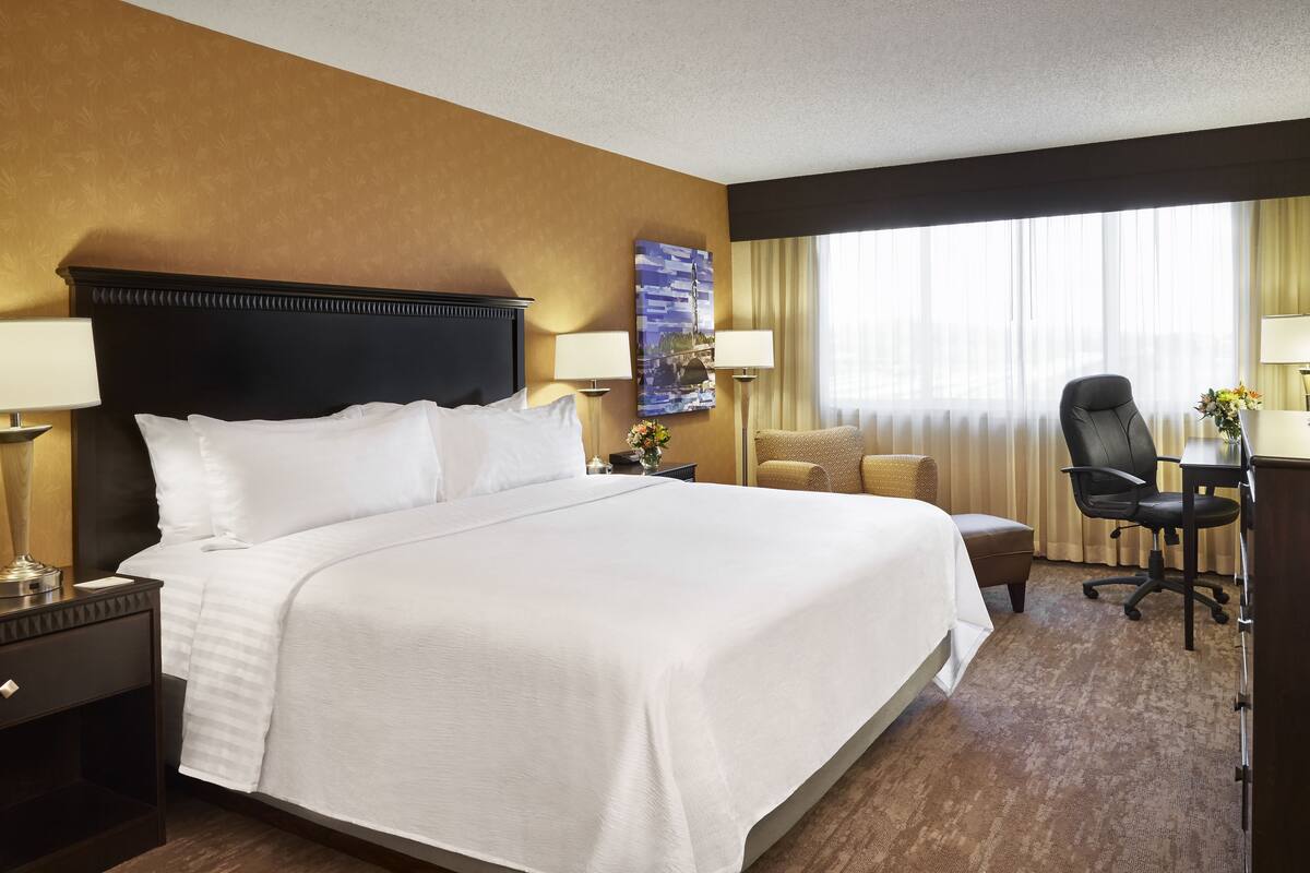 A hotel room with a large bed, a desk with a chair, a lamp, and a window with curtains.