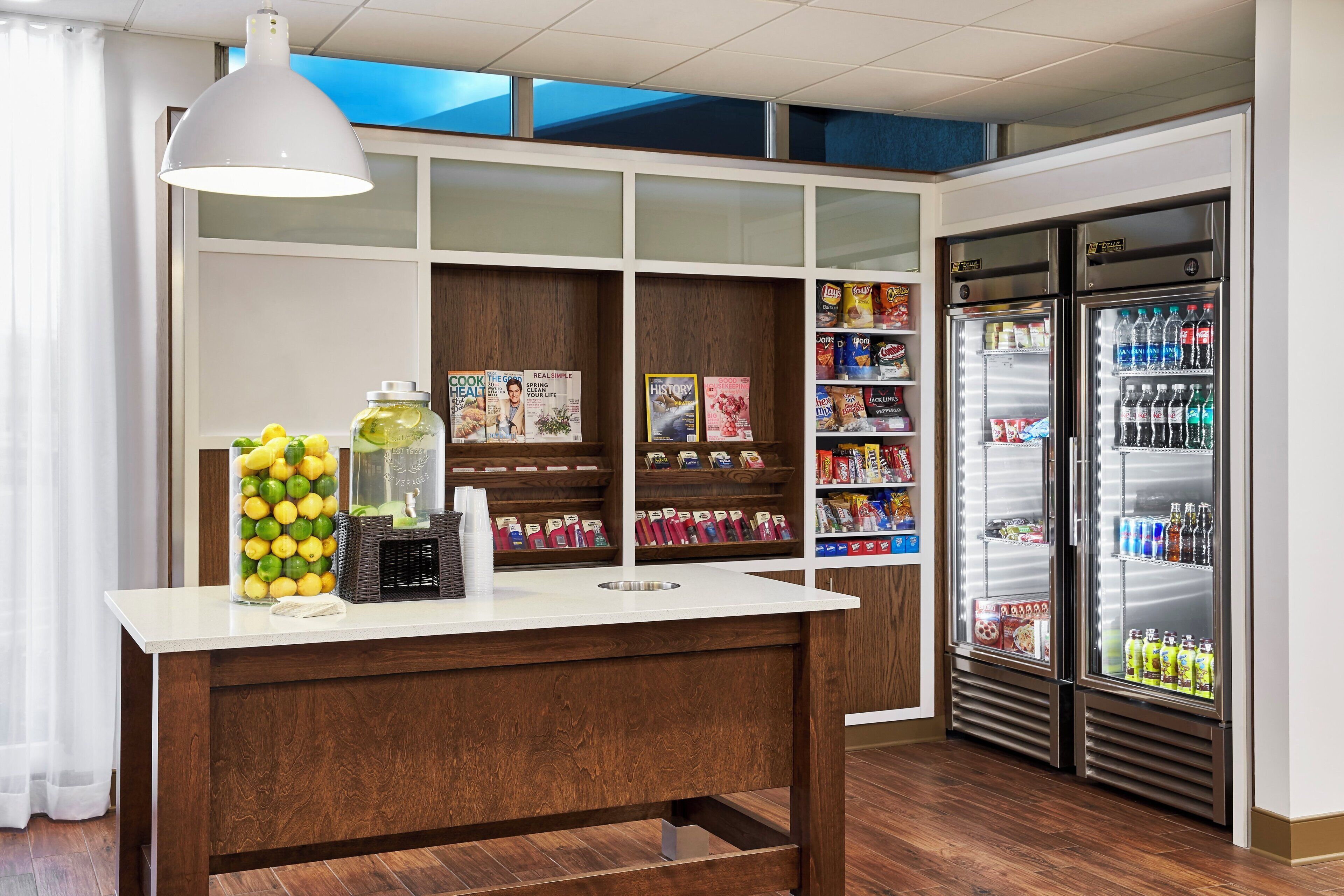 A kitchenette with a white countertop, a hanging light, a refrigerator stocked with beverages, and a shelf with magazines and snacks.