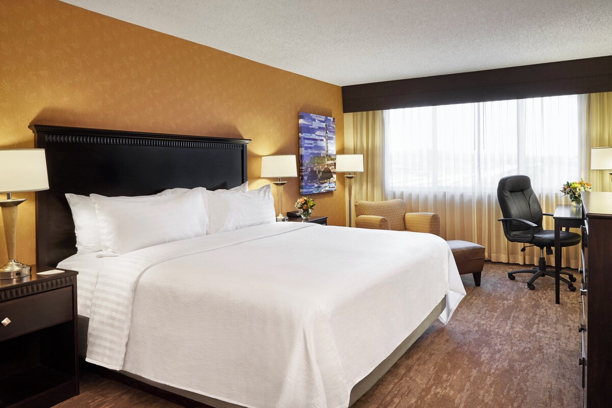 A hotel room with a large bed, a desk with a chair, a lamp, and a window with curtains.