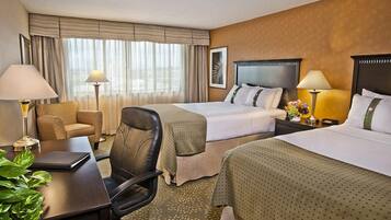 A hotel room with two beds, a desk, a chair, a lamp, and a plant.