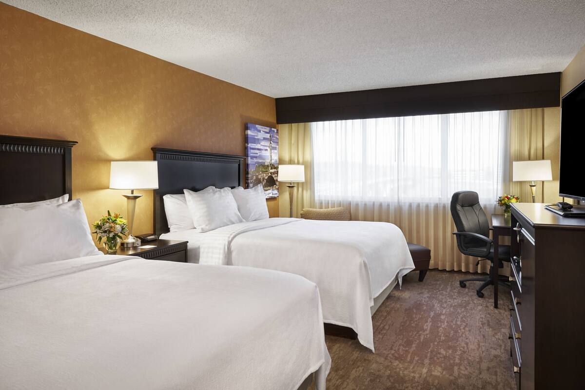 A hotel room with two beds, a desk, a chair, and a TV.
