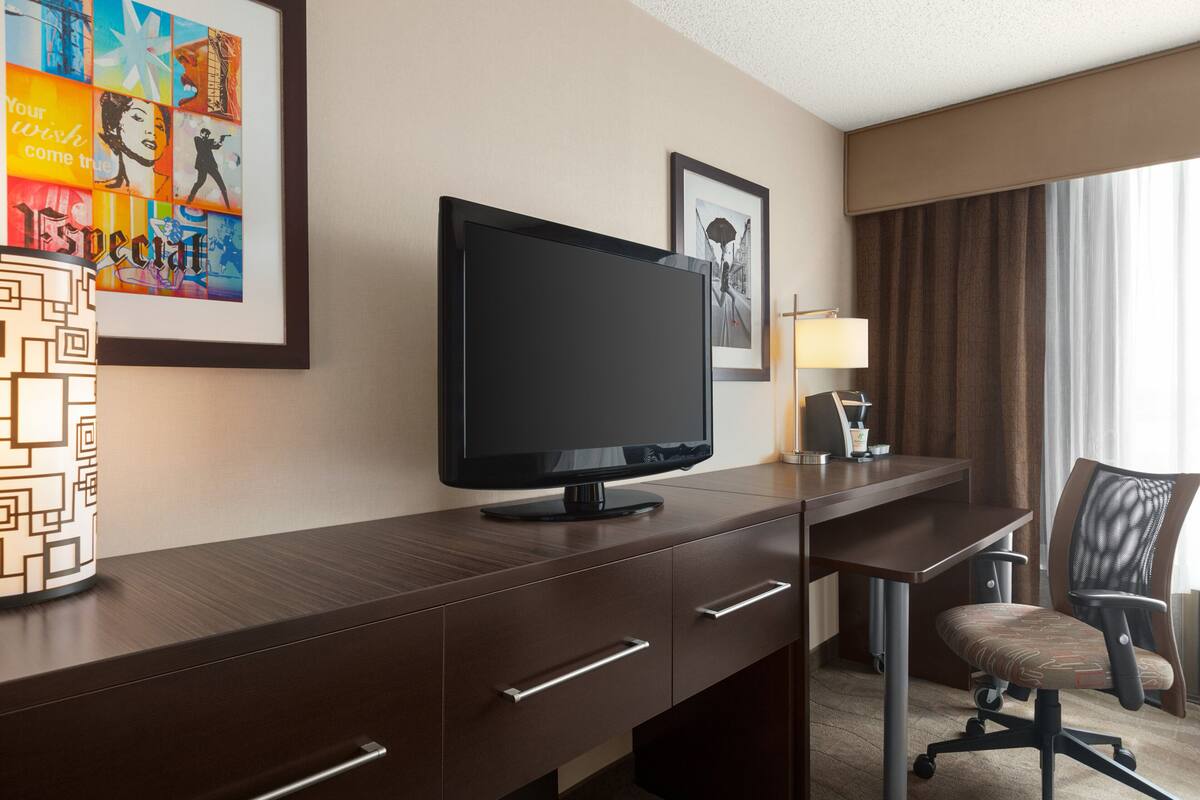 A hotel room with a desk, a television, a chair, a lamp, and artwork on the wall.