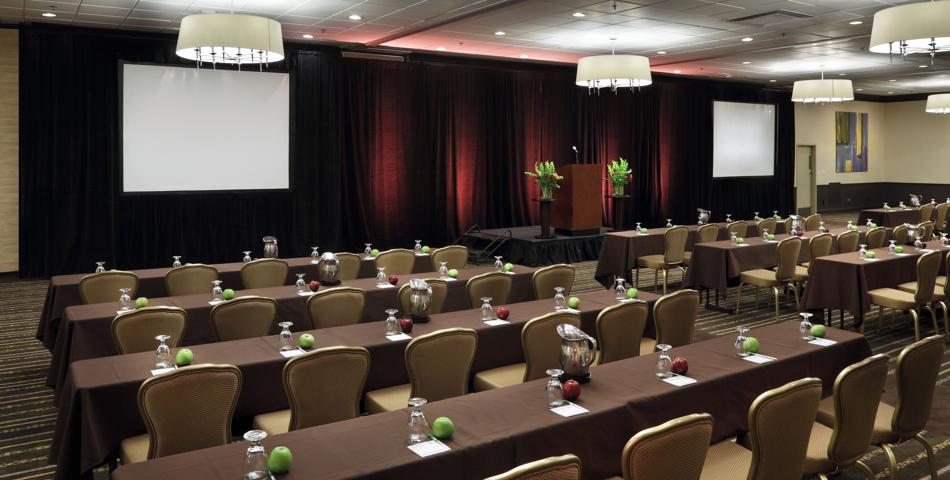 A conference room with tables set for a meeting, a stage with a podium, and a large screen.