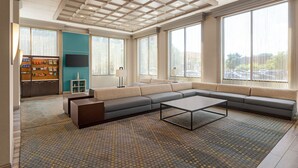 A spacious living area with a large sectional sofa, a coffee table, and a television. There are large windows providing natural light and a view of the outdoors.