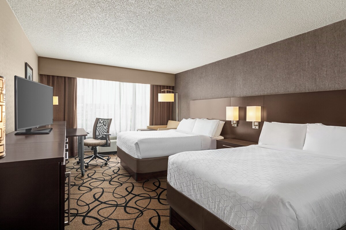 A hotel room with two beds, a desk, a chair, a TV, and a window with curtains.