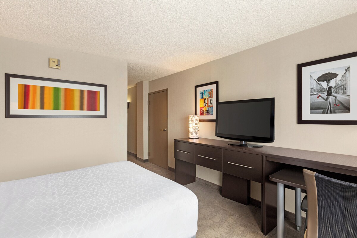A hotel room with a bed, a desk with a television, and artwork on the walls.