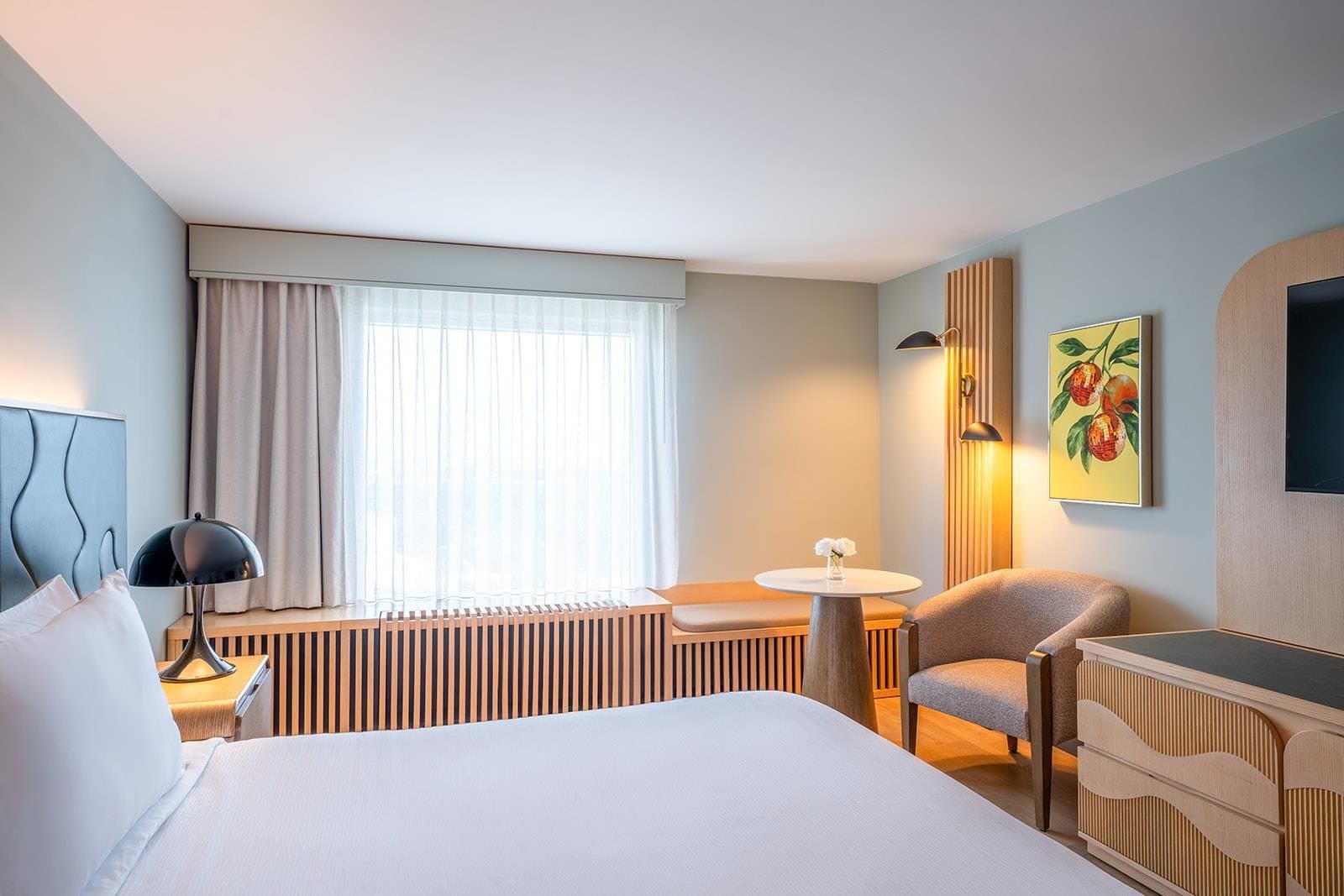 Une chambre d’hôtel comprenant un lit, un bureau, une chaise, une petite table, un radiateur, une fenêtre avec des rideaux, un téléviseur fixé au mur et un tableau.