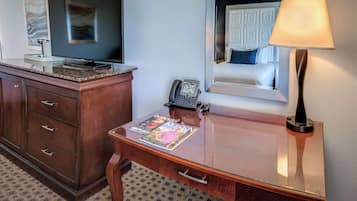 Camera d'albergo con una scrivania in legno, una TV a schermo piatto, un telefono, una rivista, una lampada e uno specchio che riflette un letto con un cuscino blu.