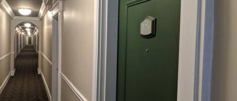 Pasillo de hotel con piso alfombrado, paredes blancas y una puerta verde con placa de número.