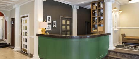 Zona de recepción con un mostrador verde, columnas blancas, una escalera de mármol y una estantería con libros y objetos decorativos.