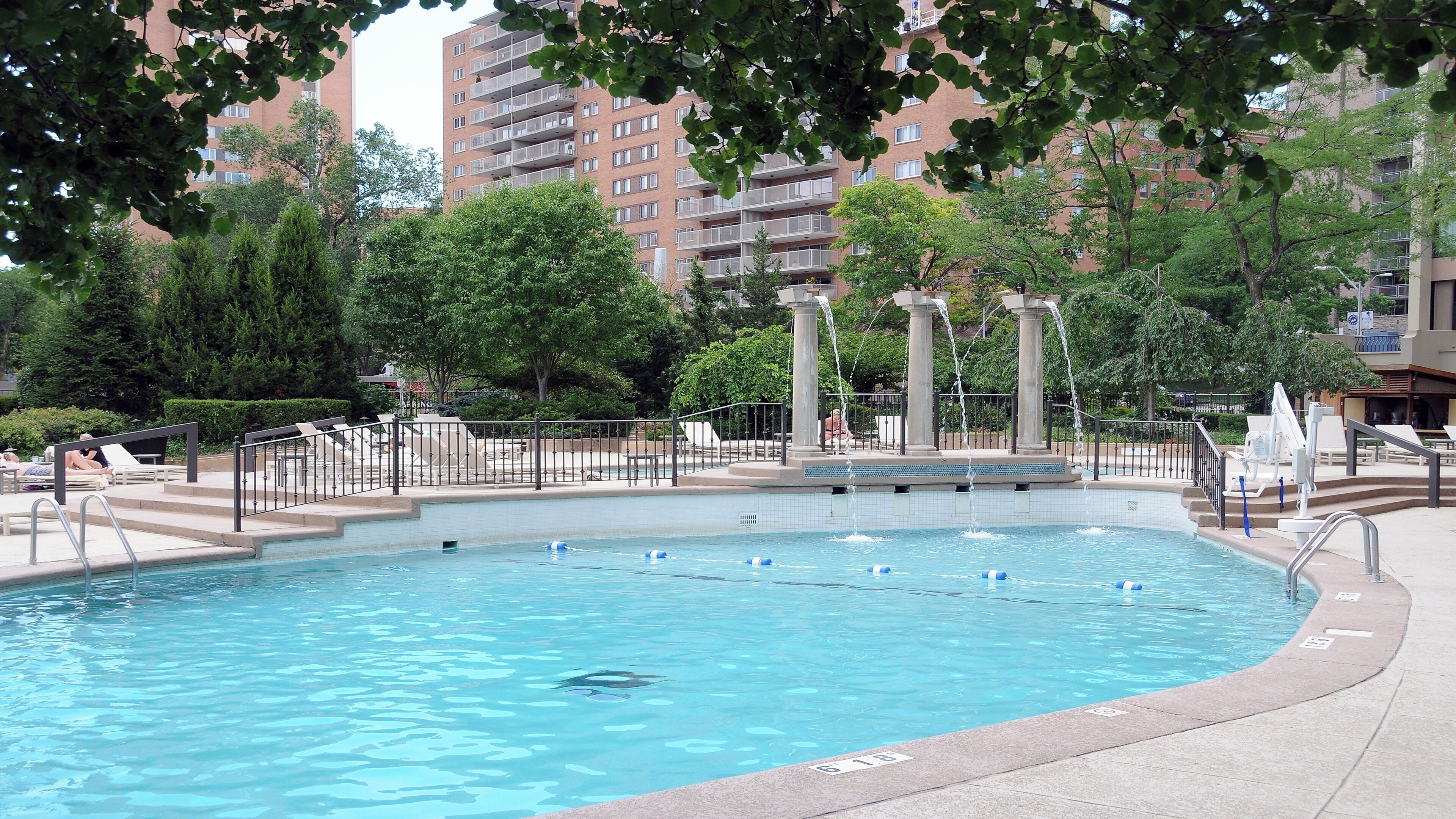 A swimming pool with a water fountain, surrounded by trees and a fence.