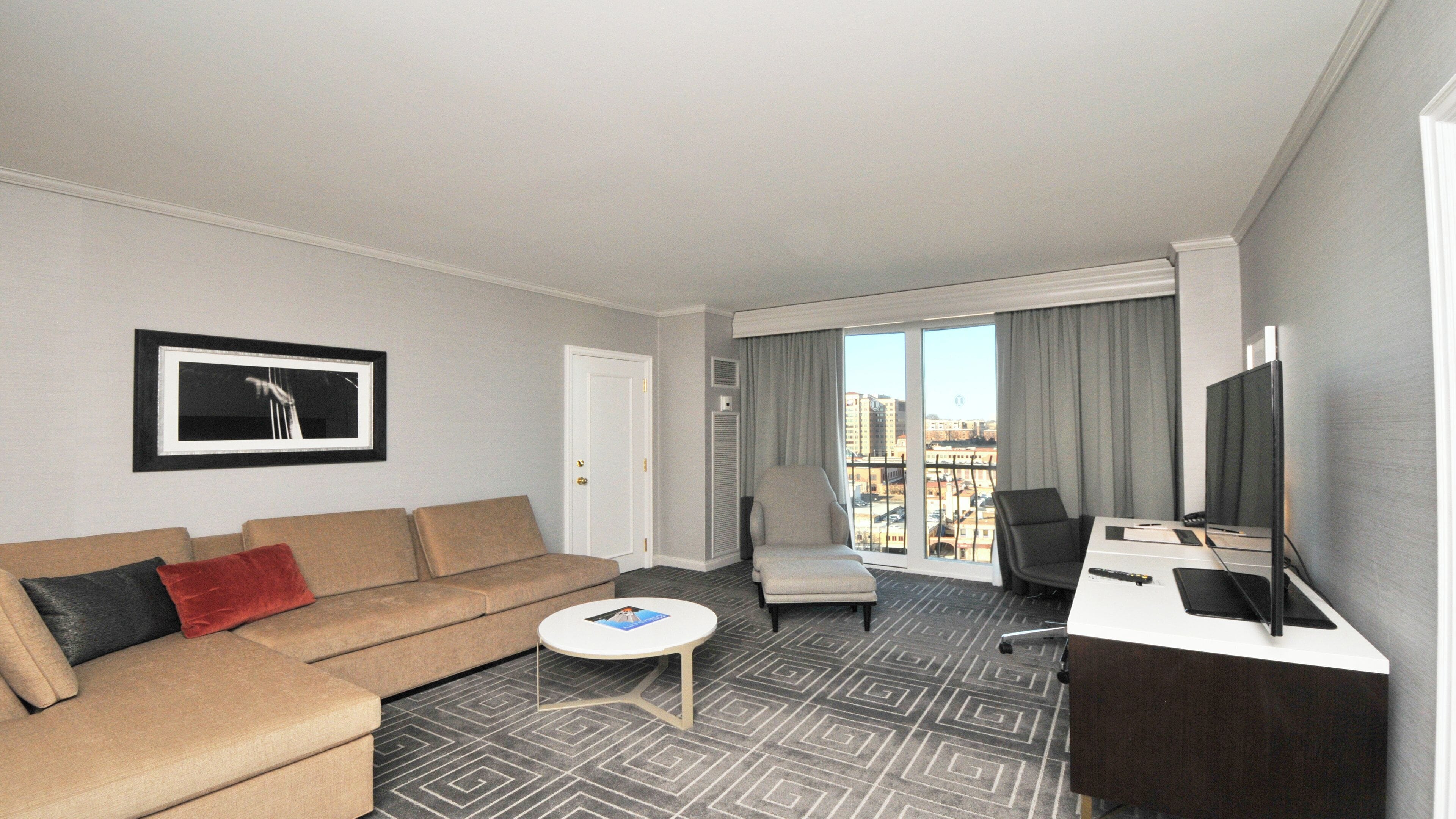 A hotel room with a sofa, armchair, desk, and a large window with a city view.