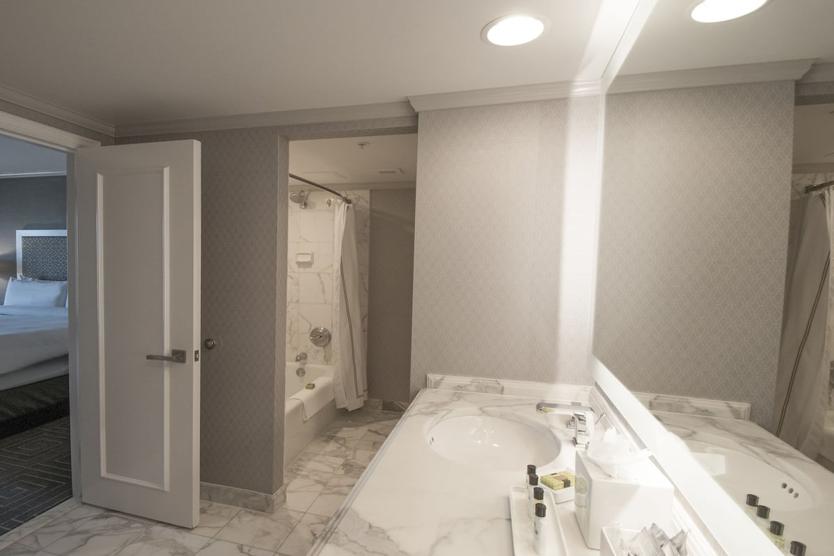 Baño de hotel con una bañera grande, zona de ducha separada y una habitación visible con cama y mesita de noche.