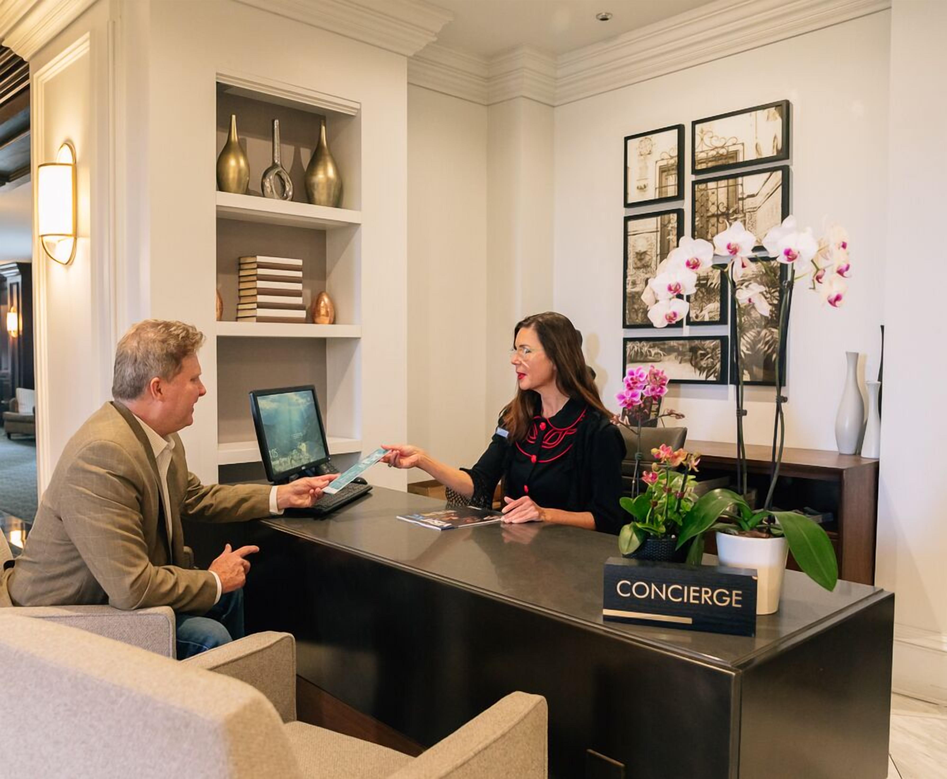 A reception desk with a concierge sign, a laptop, and a woman handing a card to a man.