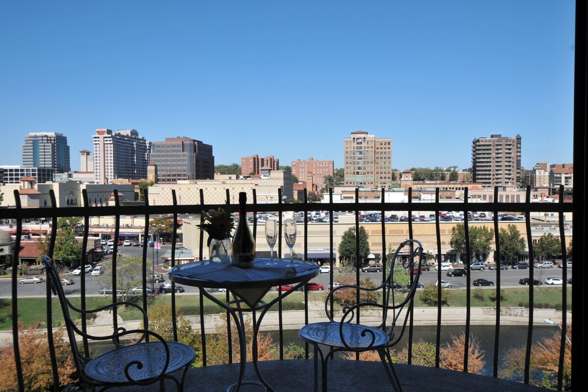 Un balcón con una mesa, dos sillas y vista a un paisaje urbano con edificios y un estacionamiento.
