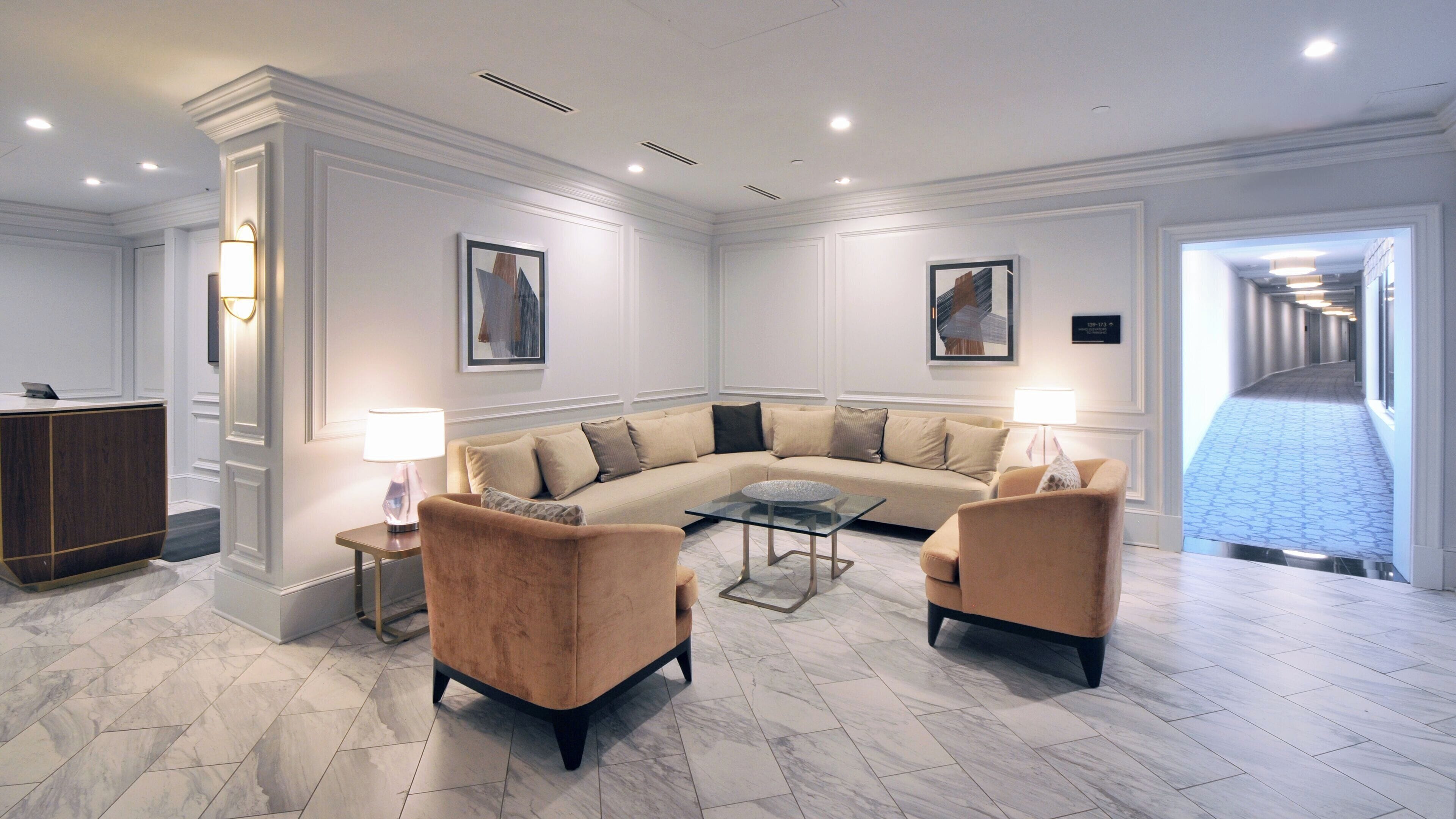 Una sala de estar moderna con un sofá seccional color beige, dos sillones, una mesa de centro de vidrio y un escritorio con una lámpara.