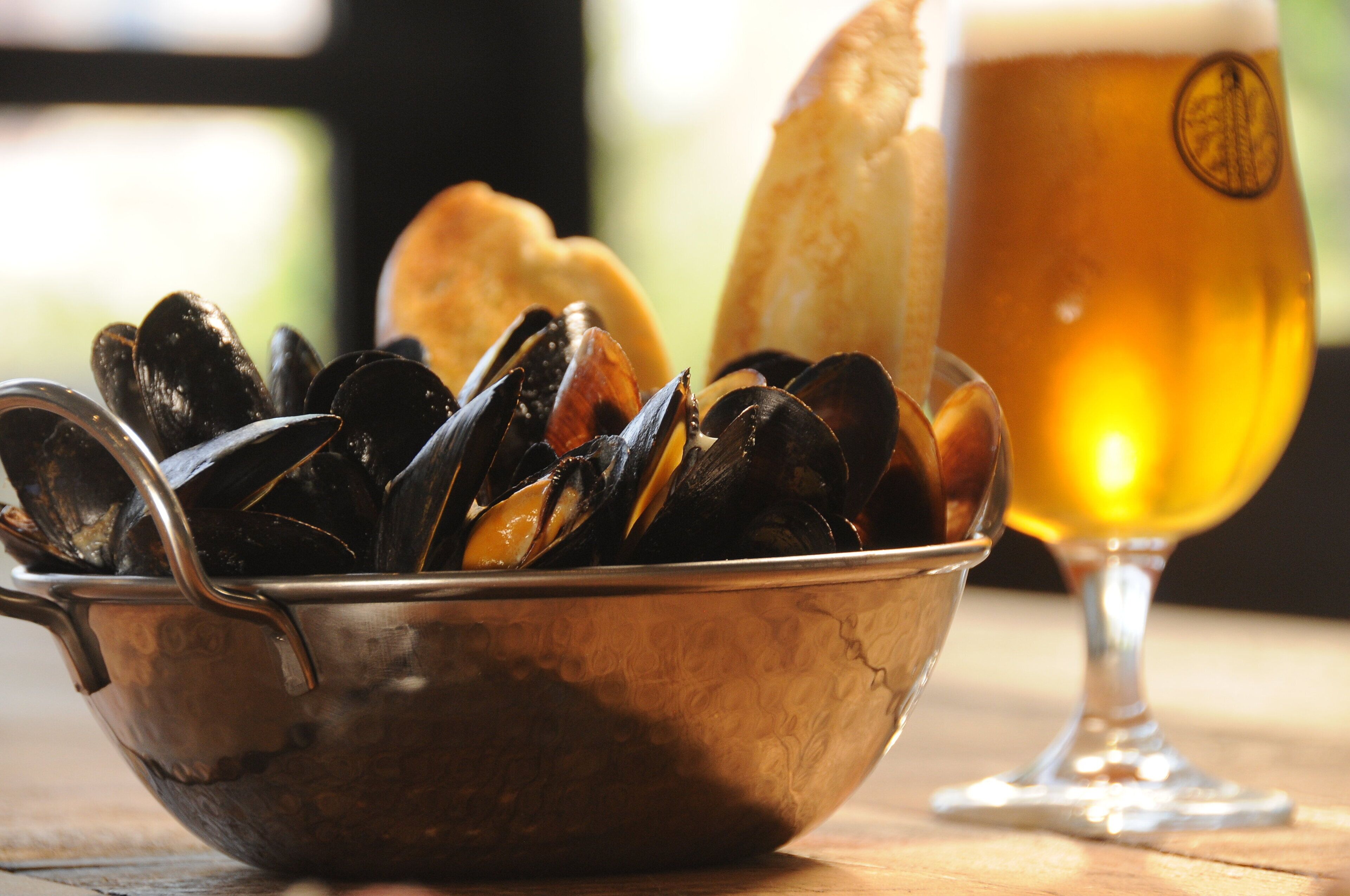 A bowl of mussels, a glass of beer, and a baguette on a wooden table.
