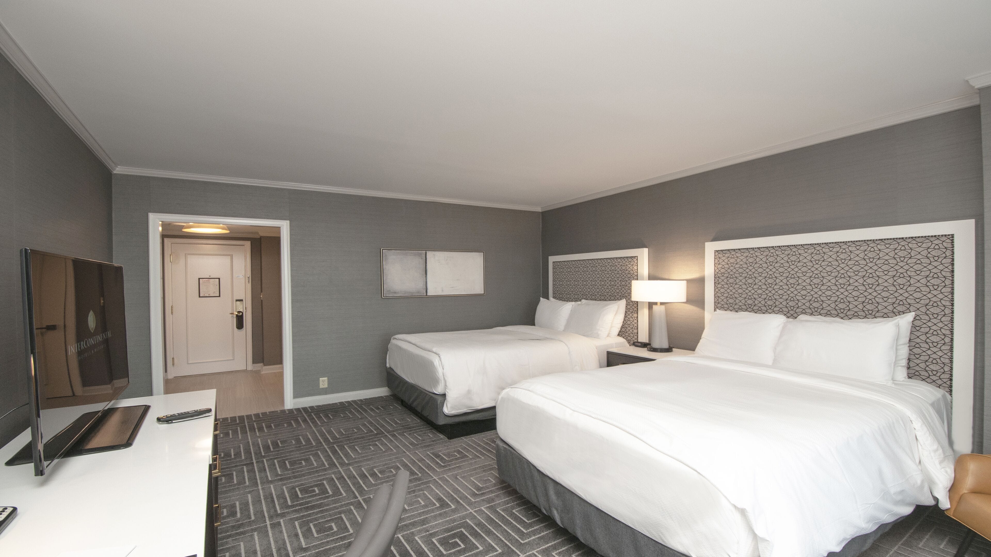 A hotel room with two beds, a desk, a TV, and a door leading to another room.