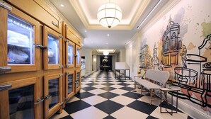 Un couloir d’hôtel avec un sol à carreaux, des armoires en bois dotées de portes en verre, un lustre et une fresque murale.