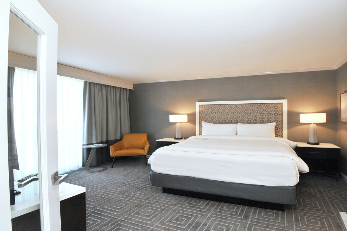 Habitación de hotel con una cama grande, dos mesitas de noche, una silla y vistas al exterior.