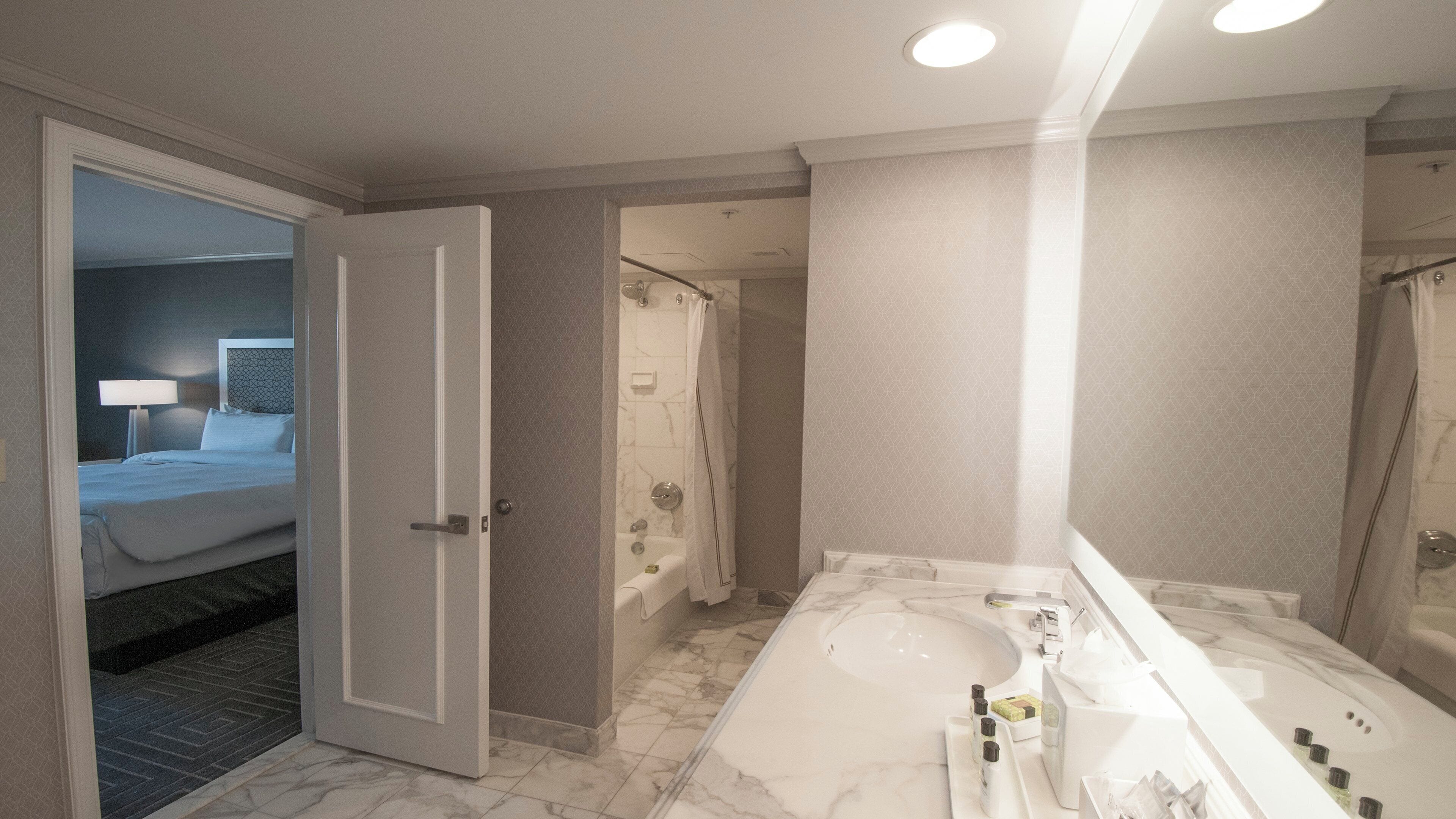 A hotel bathroom with a large bathtub, a separate shower area, and a bedroom visible through an open door.