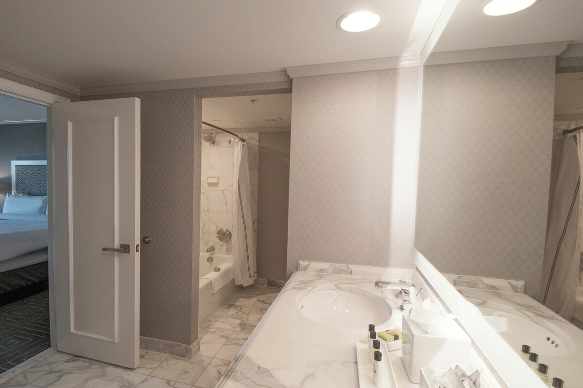 Un baño de hotel con una bañera grande, una zona de ducha separada y un dormitorio visible a través de una puerta abierta.