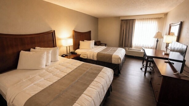 Ein Hotelzimmer mit zwei Betten, einem Schreibtisch, einem Stuhl, einer Lampe und einem großen Fenster mit Vorhängen.