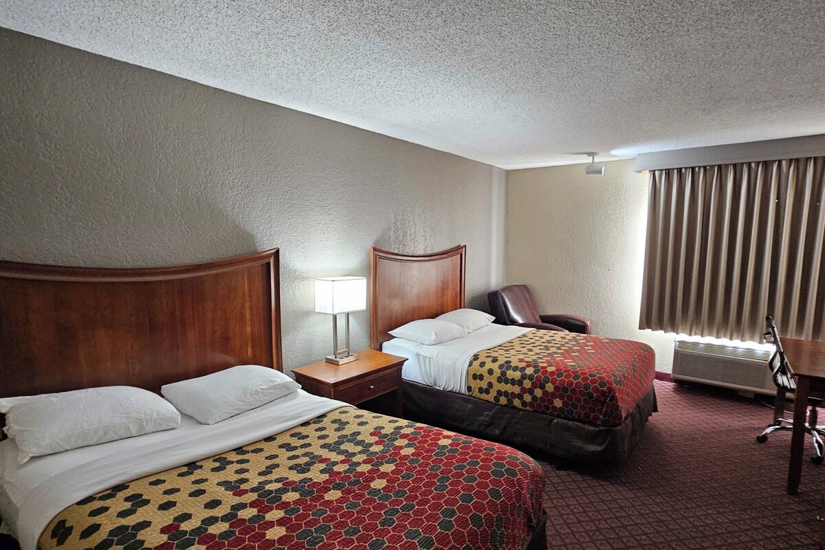 A hotel room with two beds, a desk, a chair, a lamp, and a window with curtains.