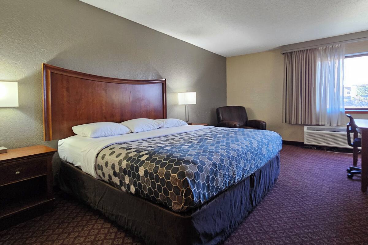 A hotel room with a large bed, a wooden headboard, a desk with a chair, a lamp, and a window with curtains.