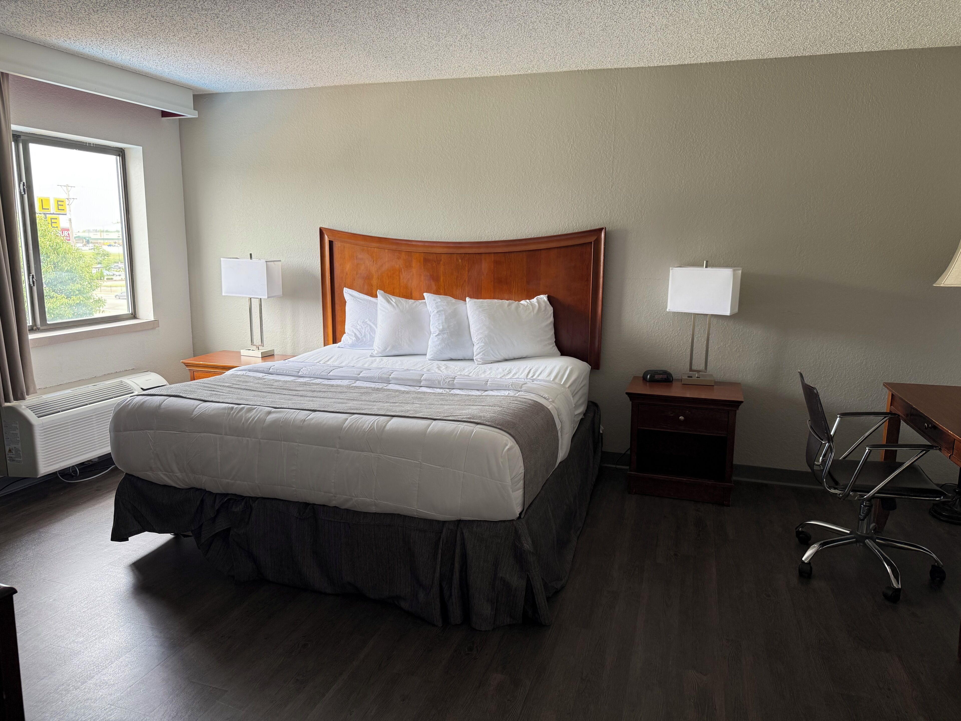 A hotel room with a large bed, two bedside tables with lamps, a desk with a chair, and a window with curtains.