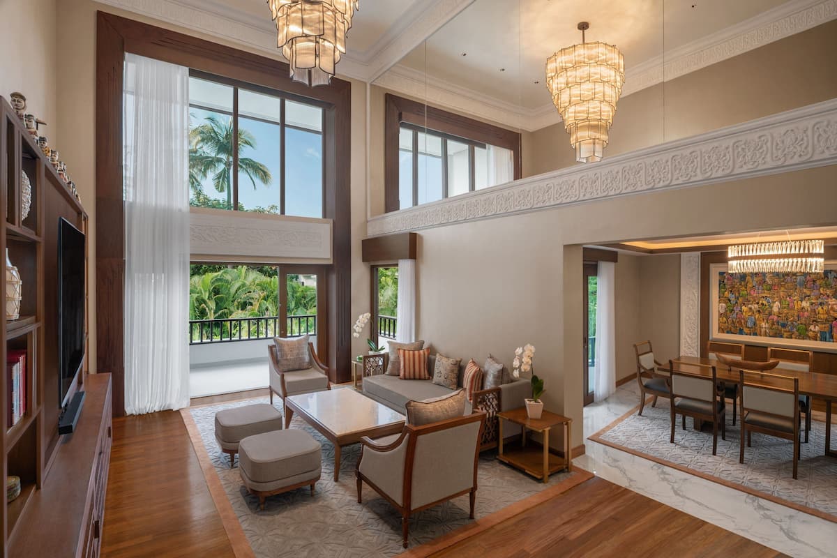 A spacious living area with a sofa set, coffee table, and dining table. There are large windows with a view of greenery outside.