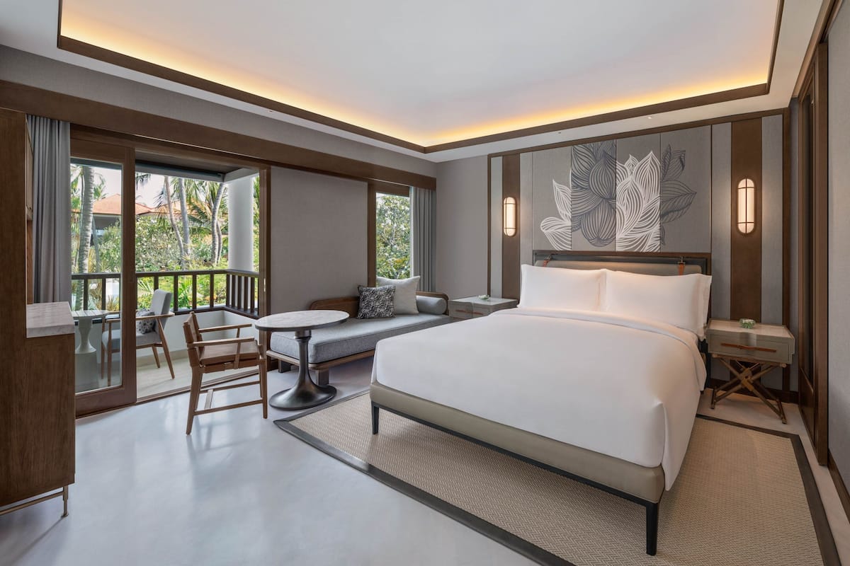 A modern hotel room with a large bed, a seating area with a table, and a balcony with a view of palm trees.