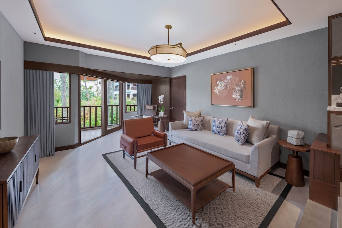 A spacious living room with a sofa, armchair, coffee table, and a large window with a view of greenery.