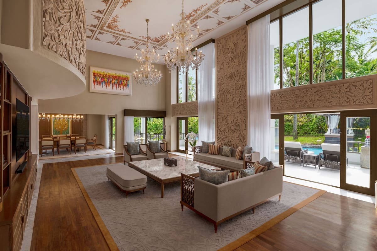 A spacious living area with a large sofa, coffee table, and a chandelier. There is a dining area with a table and chairs, and a view of the outdoors through large windows.