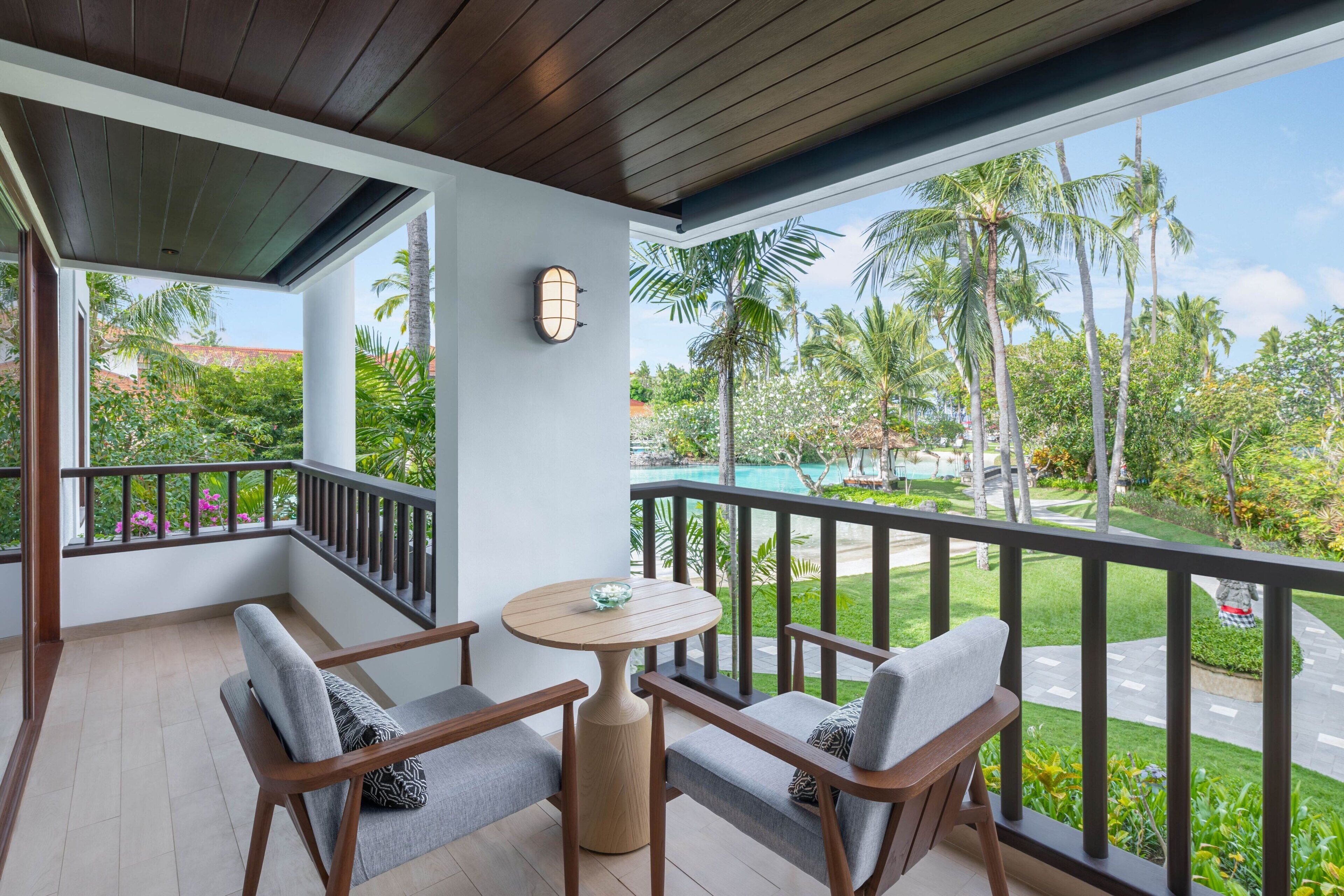A balcony with a wooden table and two chairs, a clock, and a view of a pool and greenery.