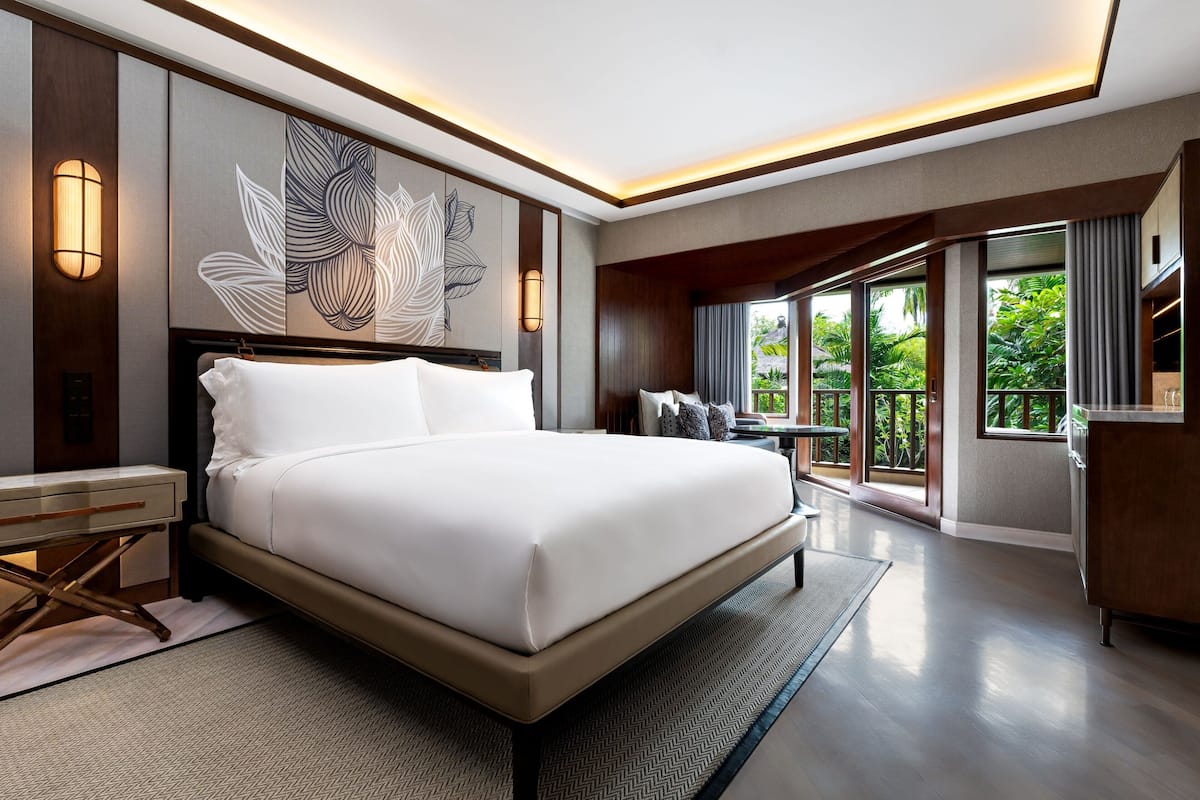 A modern hotel room with a large bed, a desk, and a view of lush greenery through the windows.