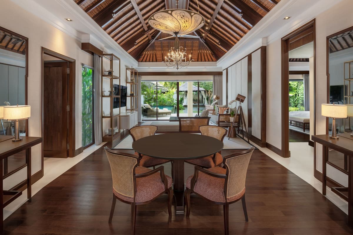 A spacious room with a large round table and six wicker chairs, a chandelier, and a view of a pool and greenery outside.