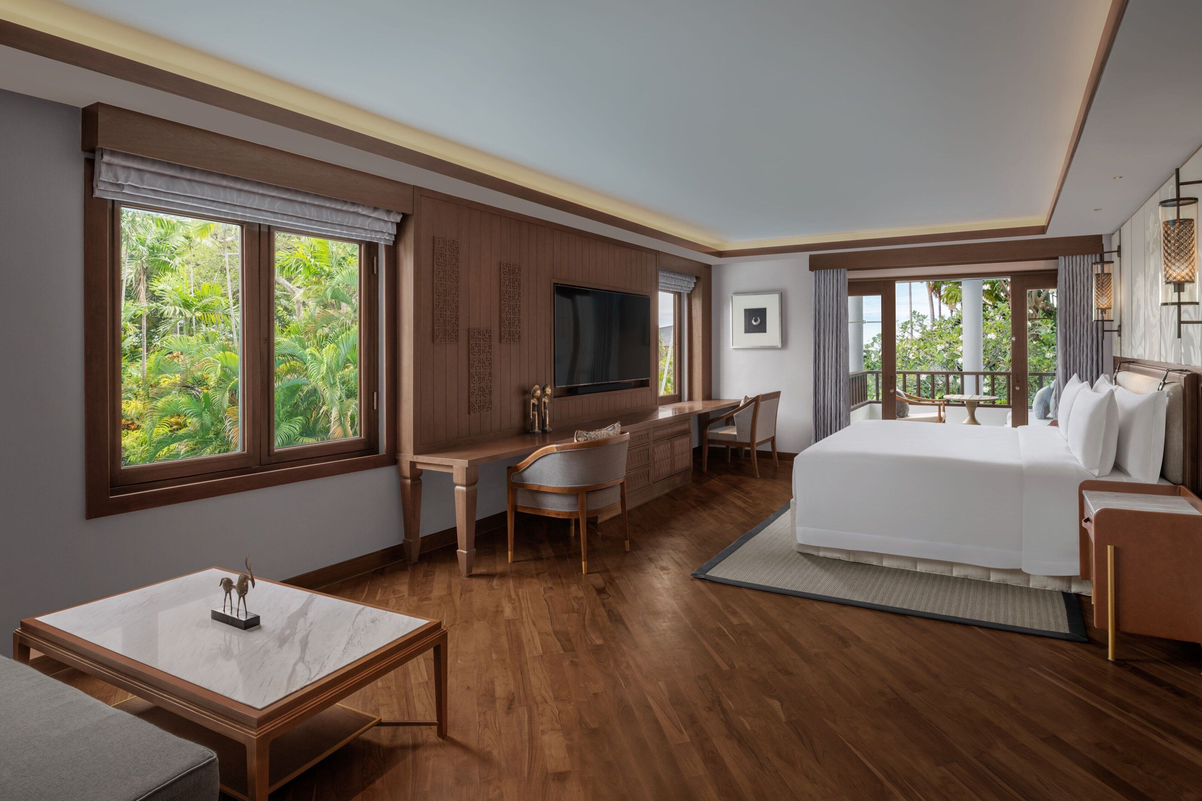 A hotel room with a large bed, a desk, a chair, a TV, and large windows with a view of greenery.