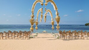 An outdoor wedding ceremony setup with a sandy beach, chairs, and decorative arches.