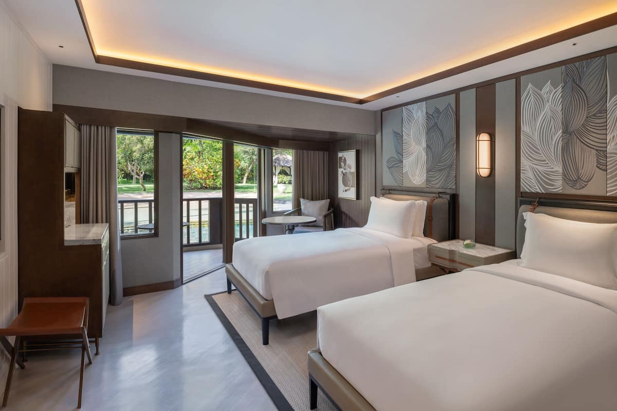 A hotel room with two beds, a small table, a chair, and a view of the outdoors.